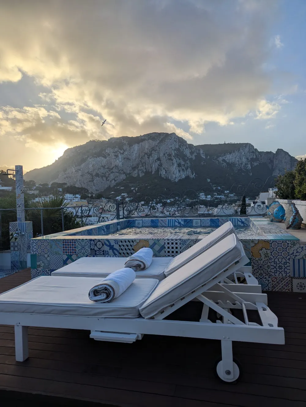 A sun lounger beside the mosaic pool at Capri Tiberio Palace at sunset, with Capri's mountain in the distance.