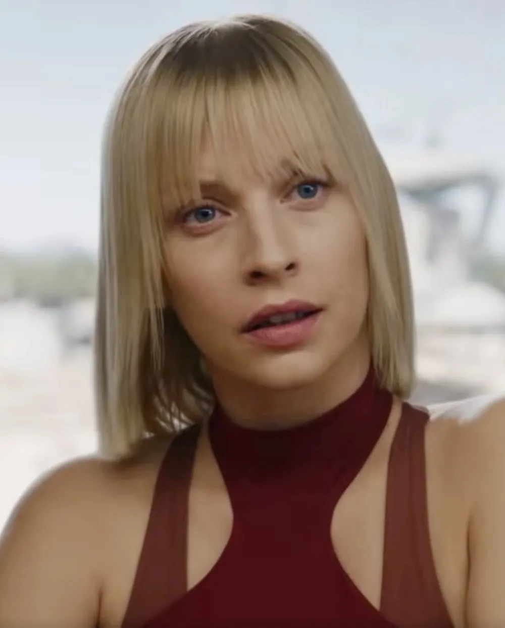 A woman with straight blonde hair and bangs is wearing a sleeveless maroon top, looking intently in a well-lit scene with a blurred outdoor background.