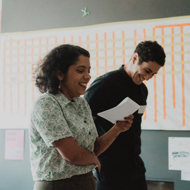 Two people smiling and reading papers in a classroom, with a chart on the wall behind them. The mood is light and collaborative.