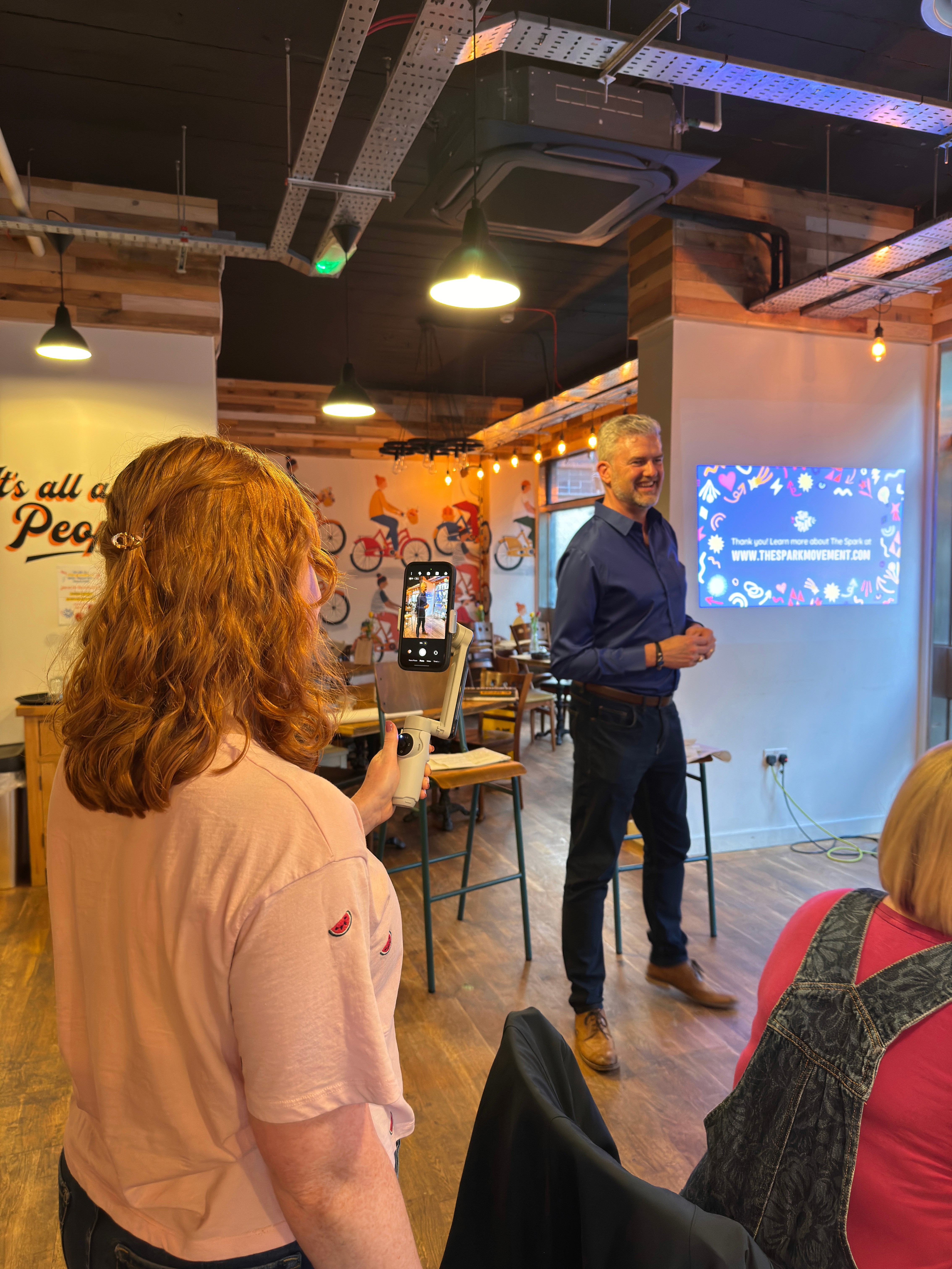 Eve filming a speaker at an indoor event using a smartphone on a handheld stabiliser.