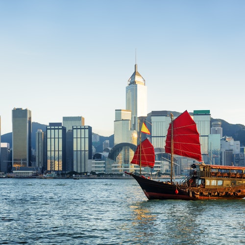 Egy hagyományos, vörös vitorlás hajó a vízen, a háttérben modern felhőkarcolók városi látképével.