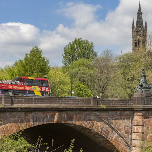 Red double-decker tour bus crossing a stone bridge with a tall church spire and trees in the background on a sunny day.