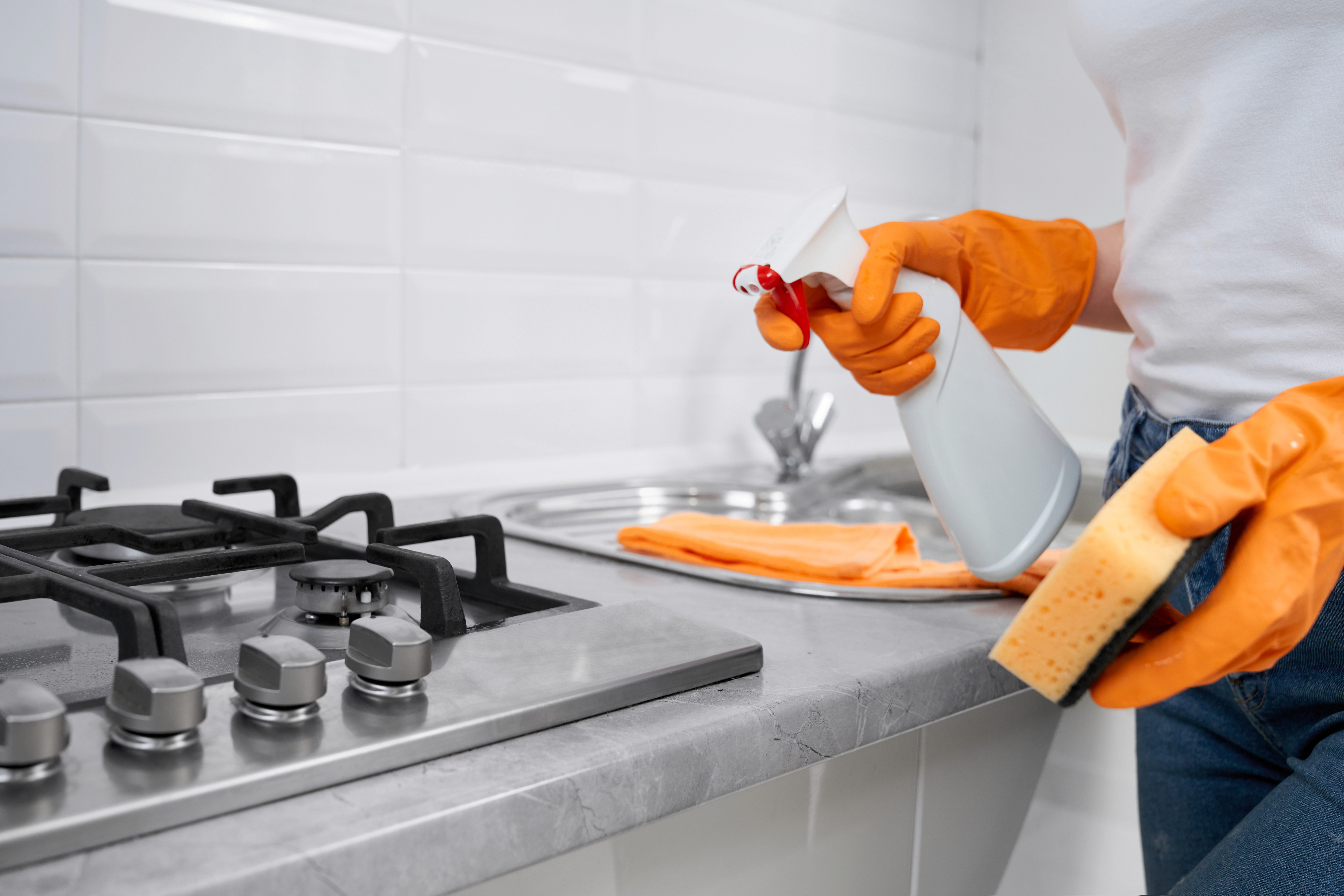 Cleaner wearing orange gloves spraying and cleaning gas stovetop in white kitchen