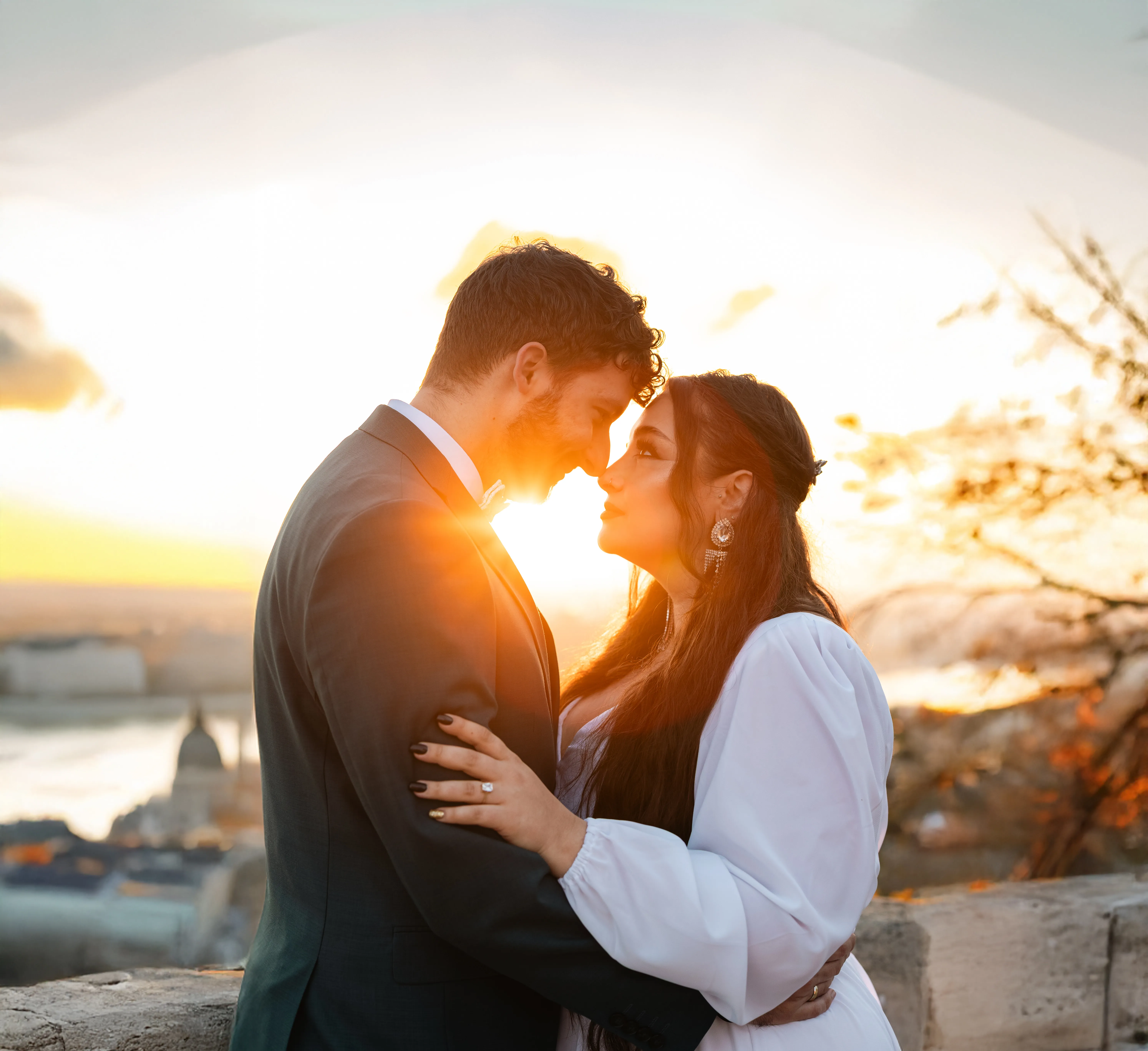 Romantikus esküvői portré napfelkelte: menyasszony fehér blúzban és vőlegény öltönyben egymást ölelve, aranyóra fény háttérvilágítással,