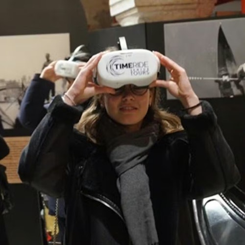 Una persona che indossa un visore per realtà virtuale con la scritta 'TimeRide' in una stanza poco illuminata, con foto esposte alle pareti.