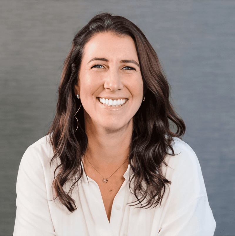 Smiling woman with long, dark hair wearing a white top, sitting against a gray background.