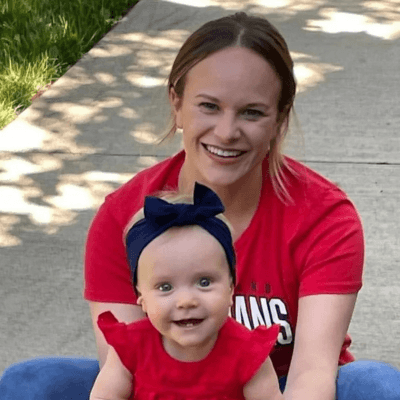 Portrait of Hillary Heaton smiling while sitting outdoors with her baby dressed in red and wearing a navy bow headband, both looking at the camera.