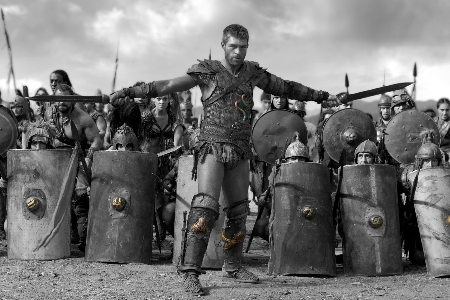 Black and white photo of a large group of gladiators holding long shiields in atraining session. In front of them Spartacus holding 2 swords  commanding their attention.