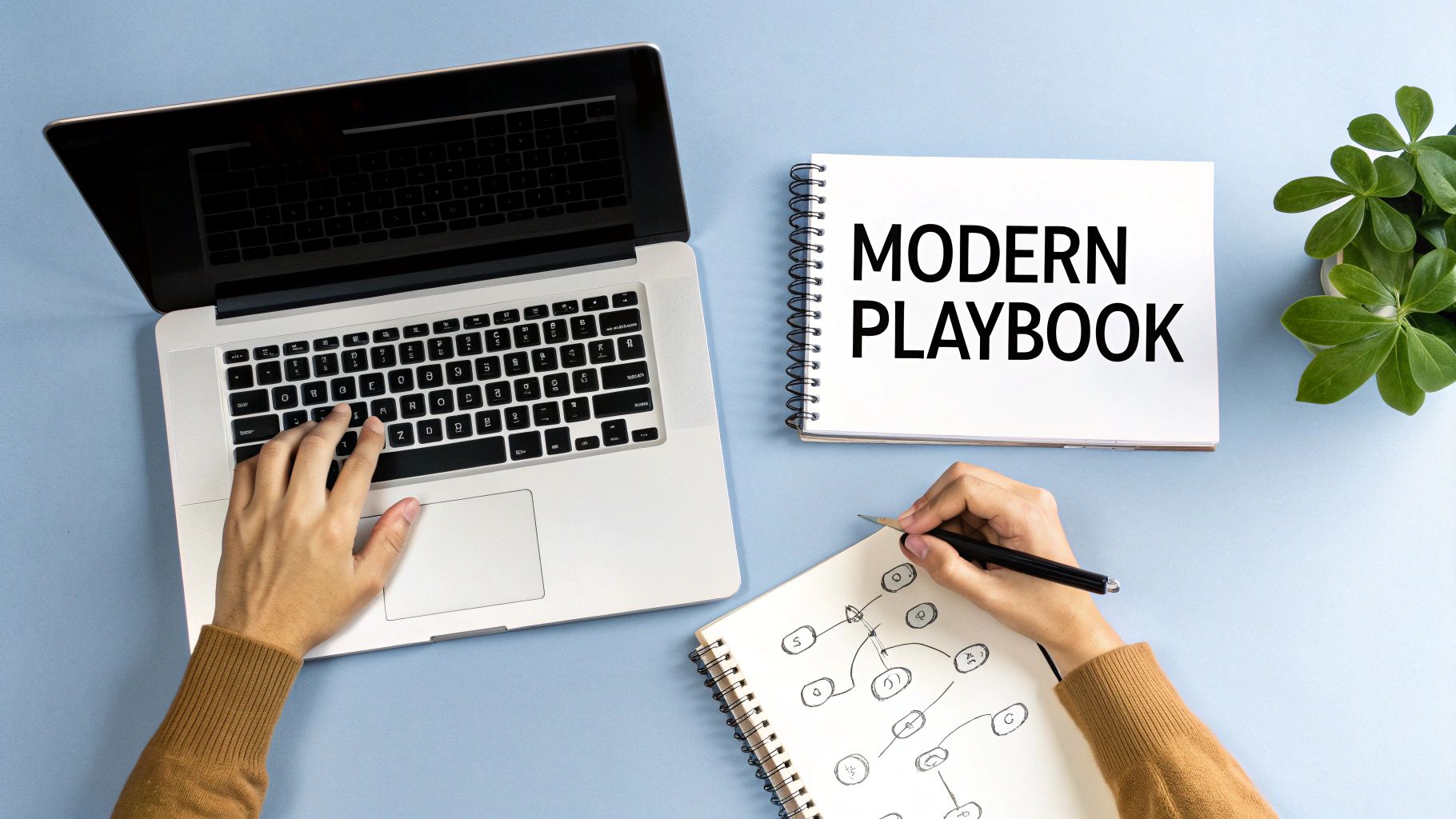 Overhead shot of a person working on a laptop and writing in a notebook labeled 'MODERN PLAYBOOK'.