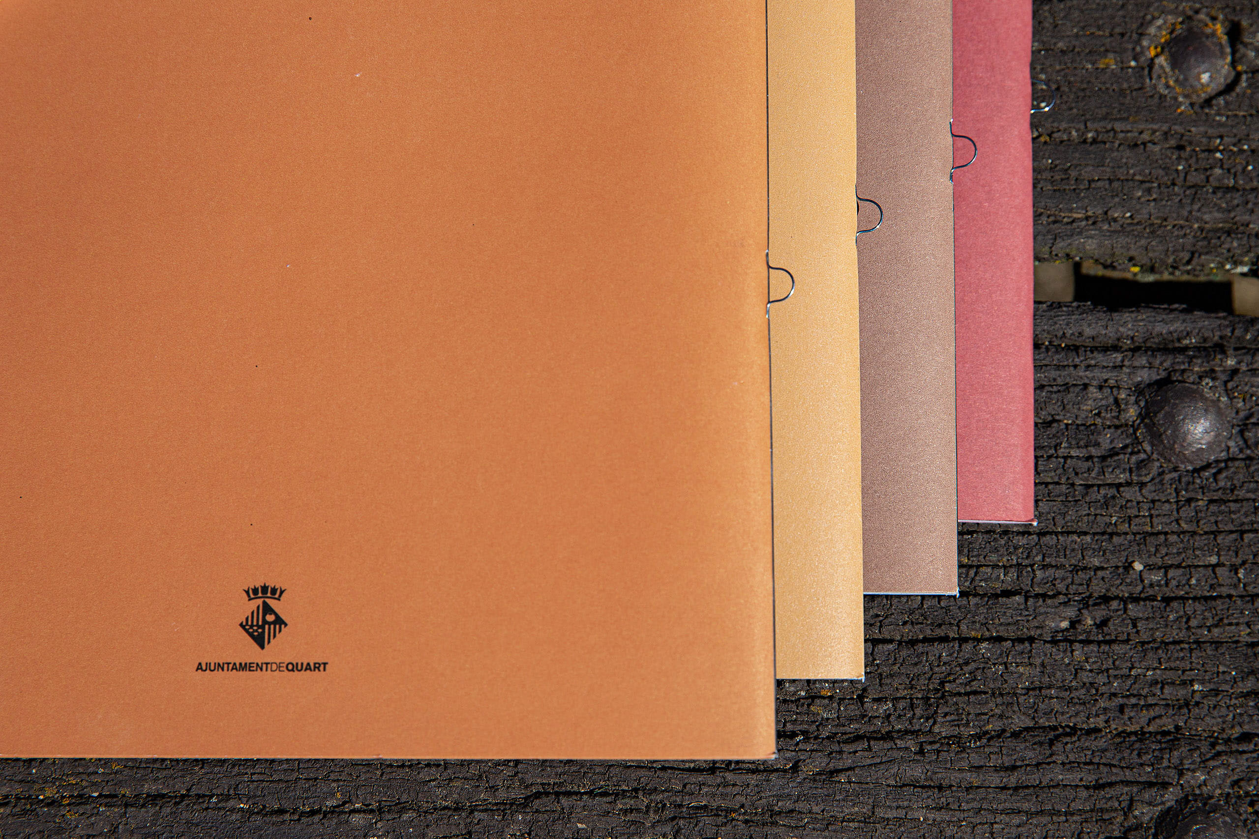 Ochre back cover with Ajuntament de Quart logo on a dark background showing stacked magazine pages, with metal spiral bindings, highlighting variations of warm earthy colors.