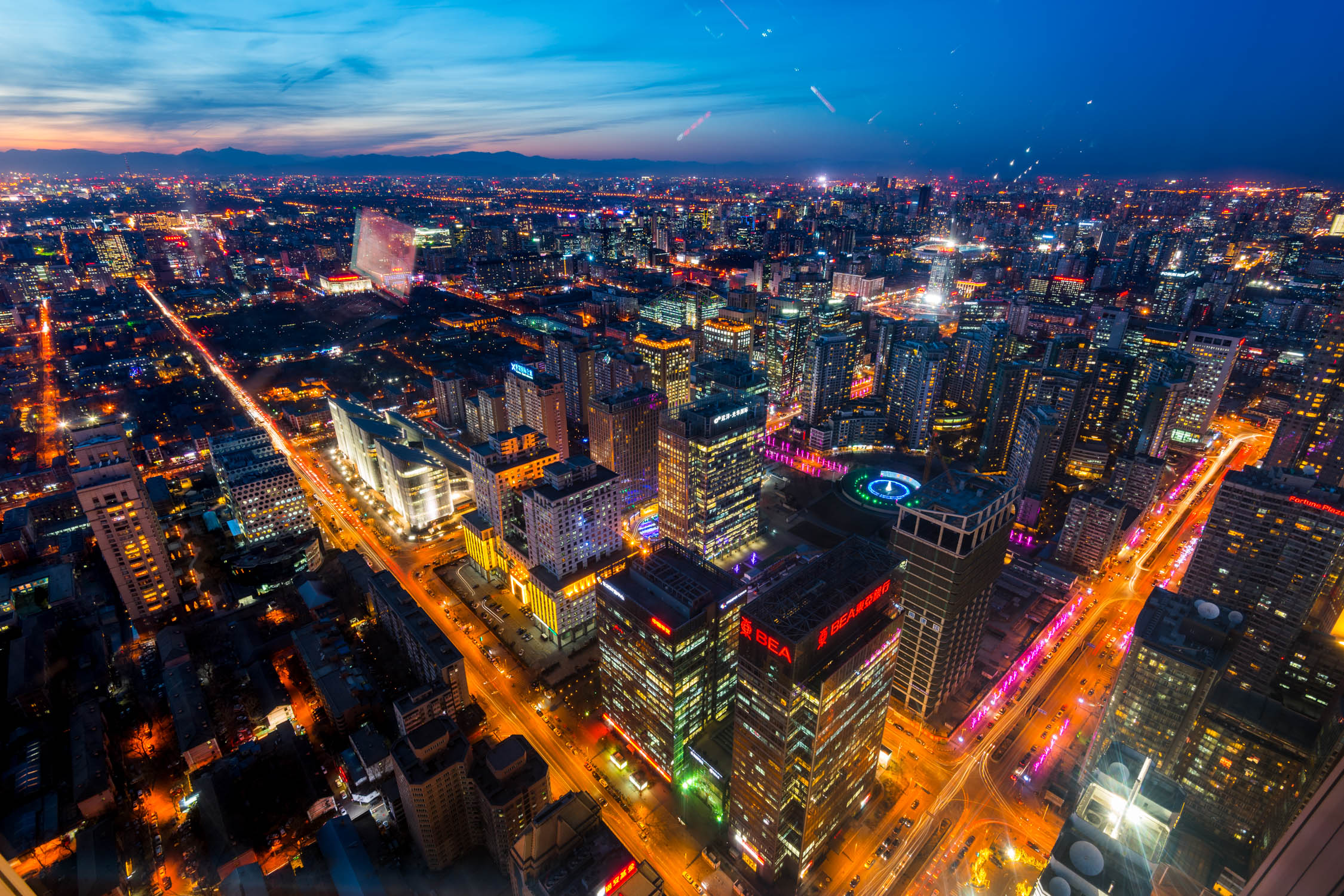 Aerial night view of a dense city skyline with glowing orange streetlights and illuminated skyscrapers under a blue sky.