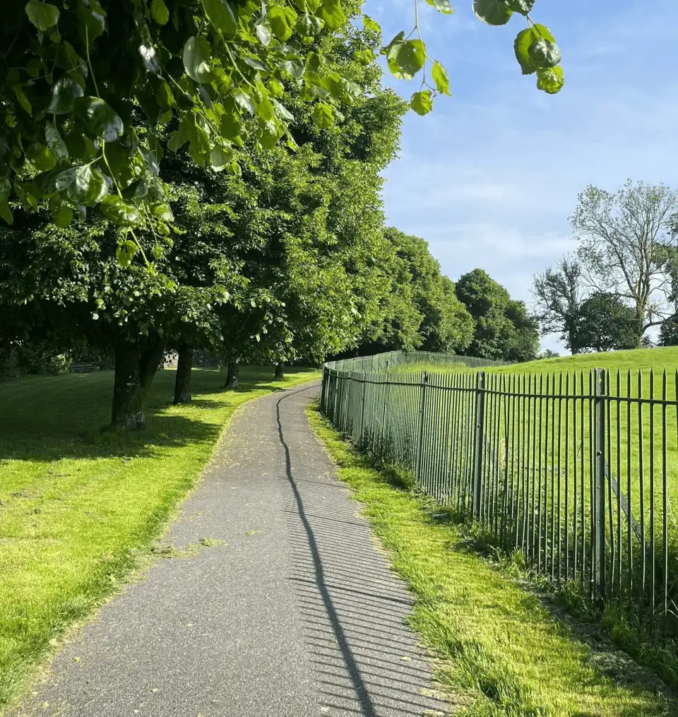 A tree-lined tarmac path through a sunlit green park in Llandeilo, bordered by a green railing fence