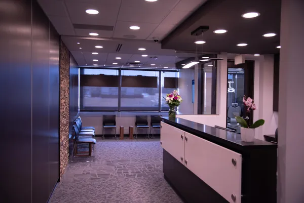Modern dental clinic reception area with a sleek black and white front desk, flower arrangements, and a row of chairs along the window wall.