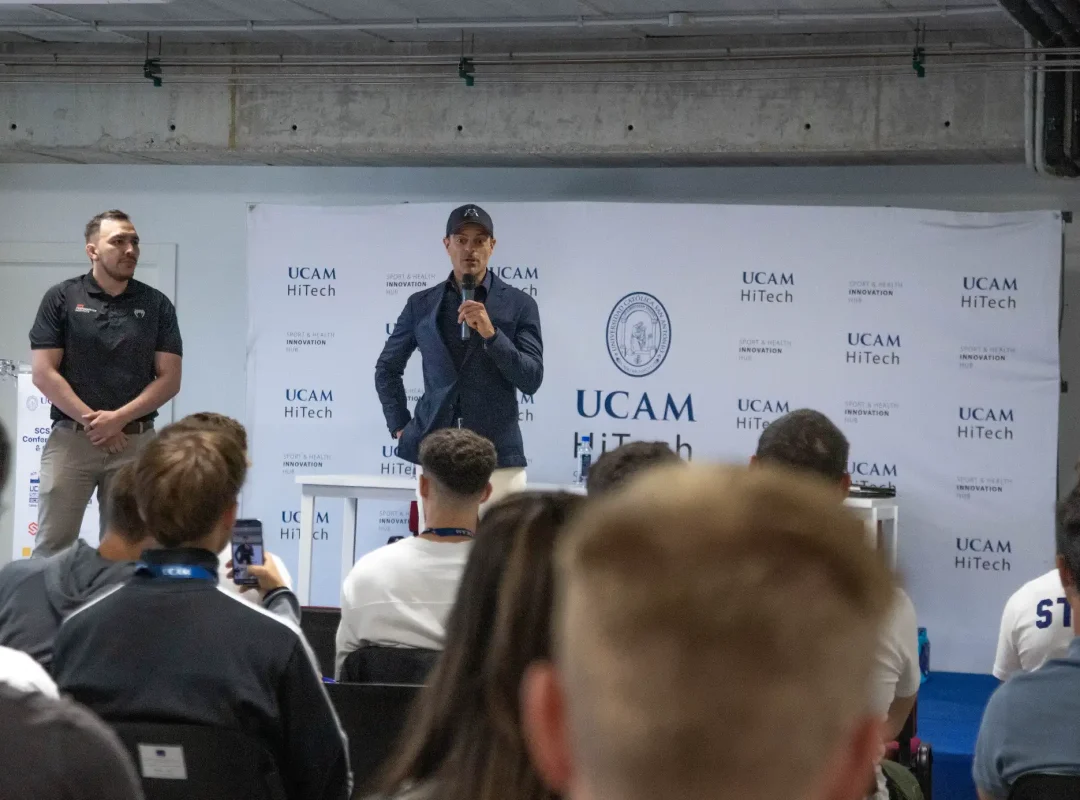 A speaker stands at a podium addressing an audience in a conference room decorated with banners featuring the European UCAM University logo and the UCAM HiTech branding.