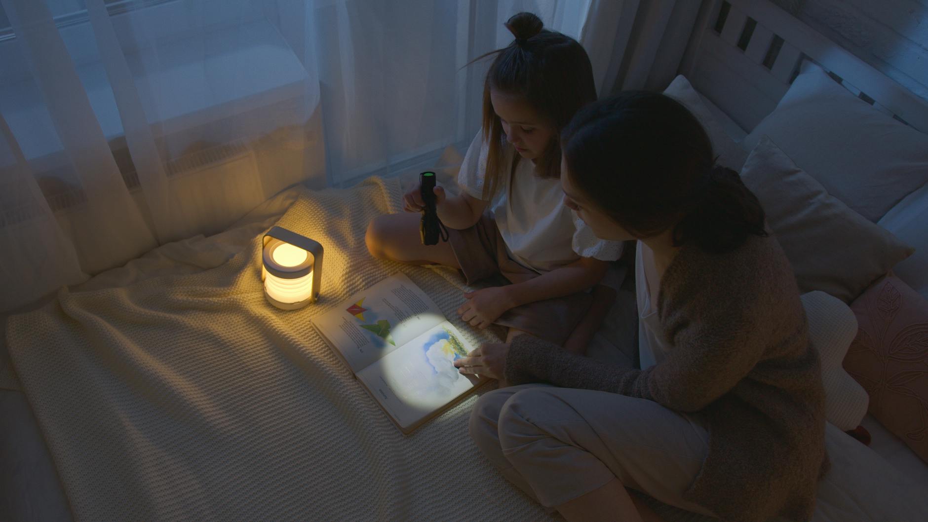 A father and child cuddled under a blanket reading a peaceful story by the glow of a bedside lamp.