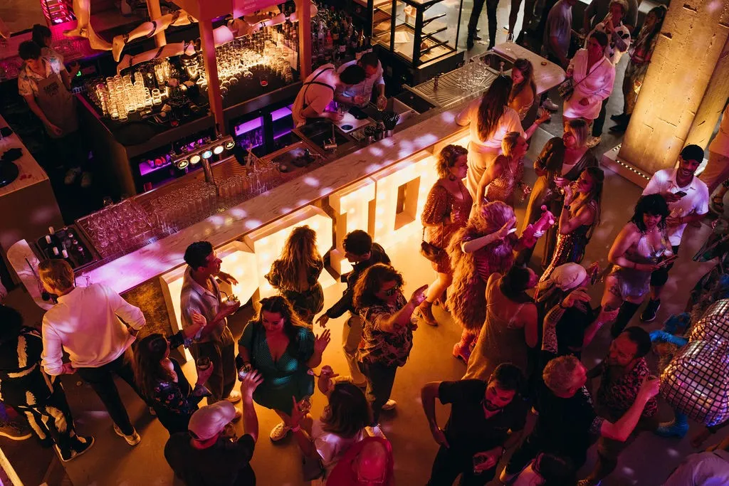 A vibrant night scene in a crowded venue with people socializing and a DJ booth in the background.