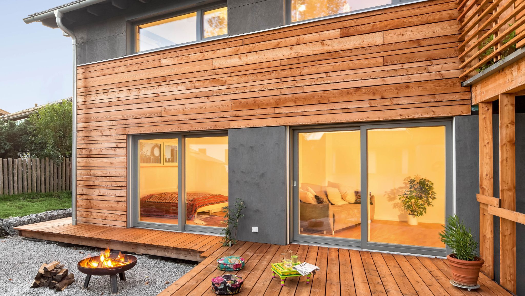 Holzverkleidete Hauswand mit großen Fenstern, Holzbalkon und Terrasse mit Feuerstelle und Sitzmöbeln.