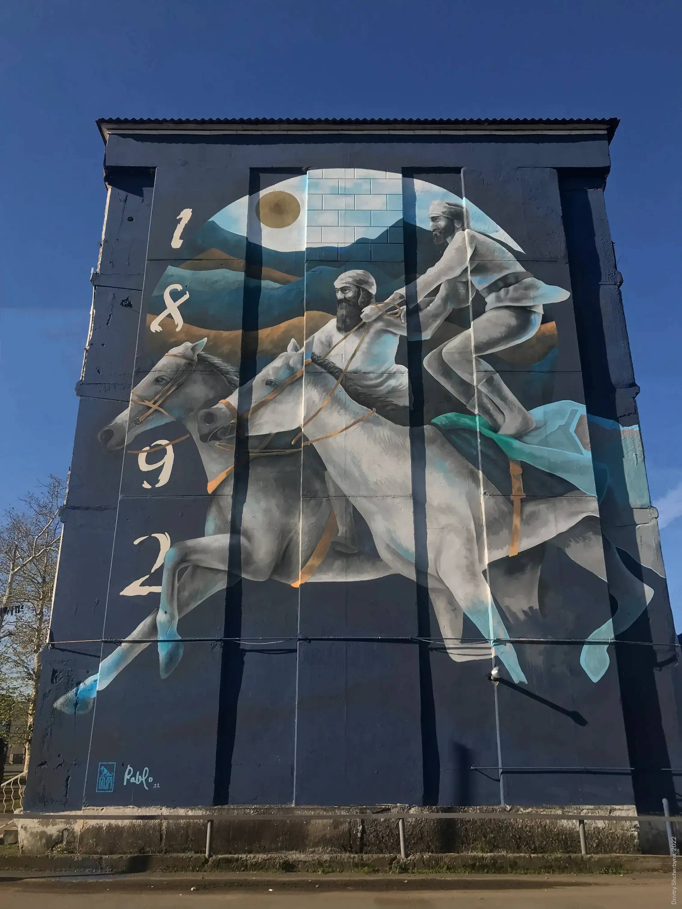 A mural on a dark blue building wall depicts two bearded horsemen in traditional Caucasian dress, galloping on grey horses, before a circular backdrop of mountains and sky.