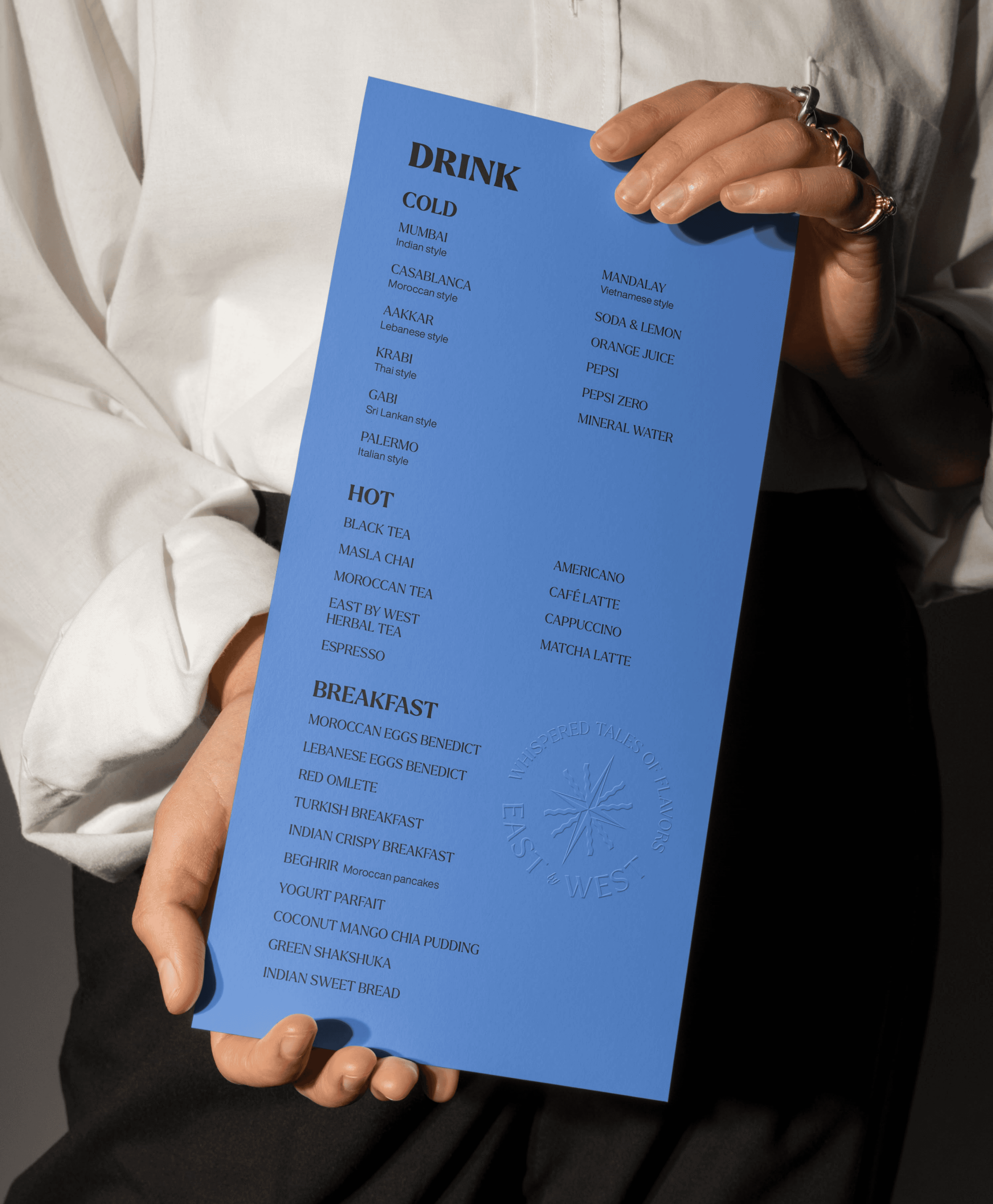 Hand holding East by West restaurant blue drinks menu listing iced lattes, teas, hot coffees, and breakfast mocktails with star emblem watermark.
