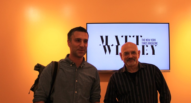 portrait of two men with a tv in they back on an orange wall