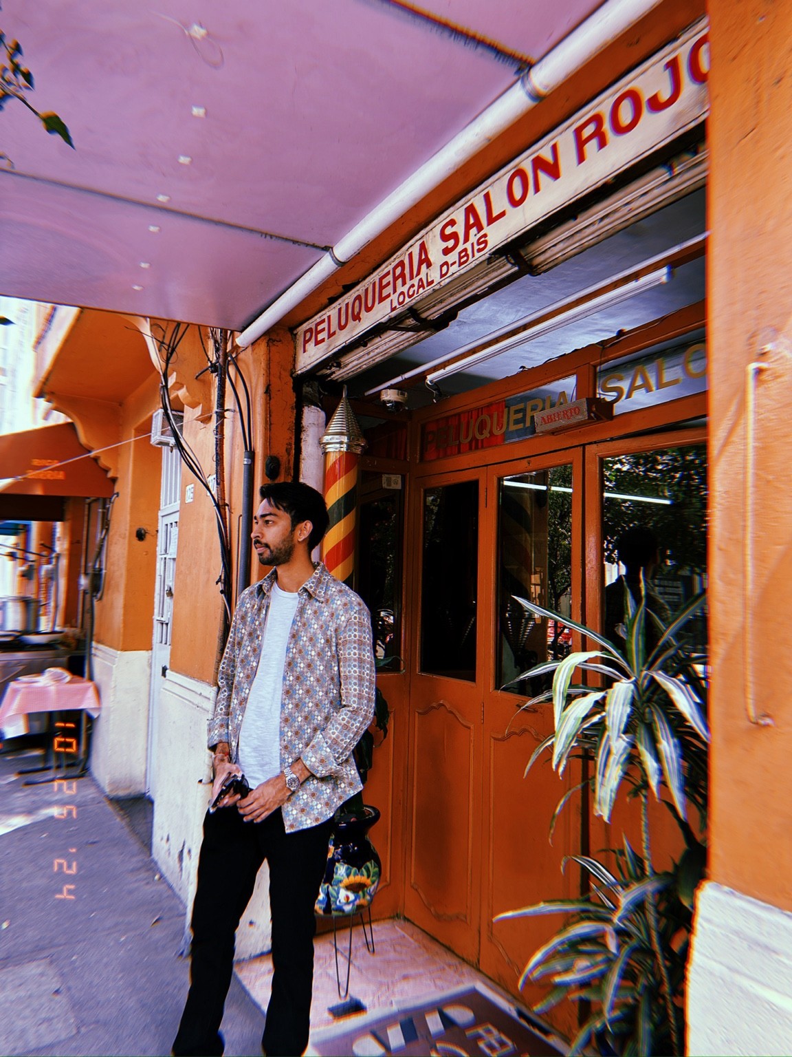 A man in a patterned shirt stands outside an orange barber shop beneath the Peluqueria Salon Rojo sign.