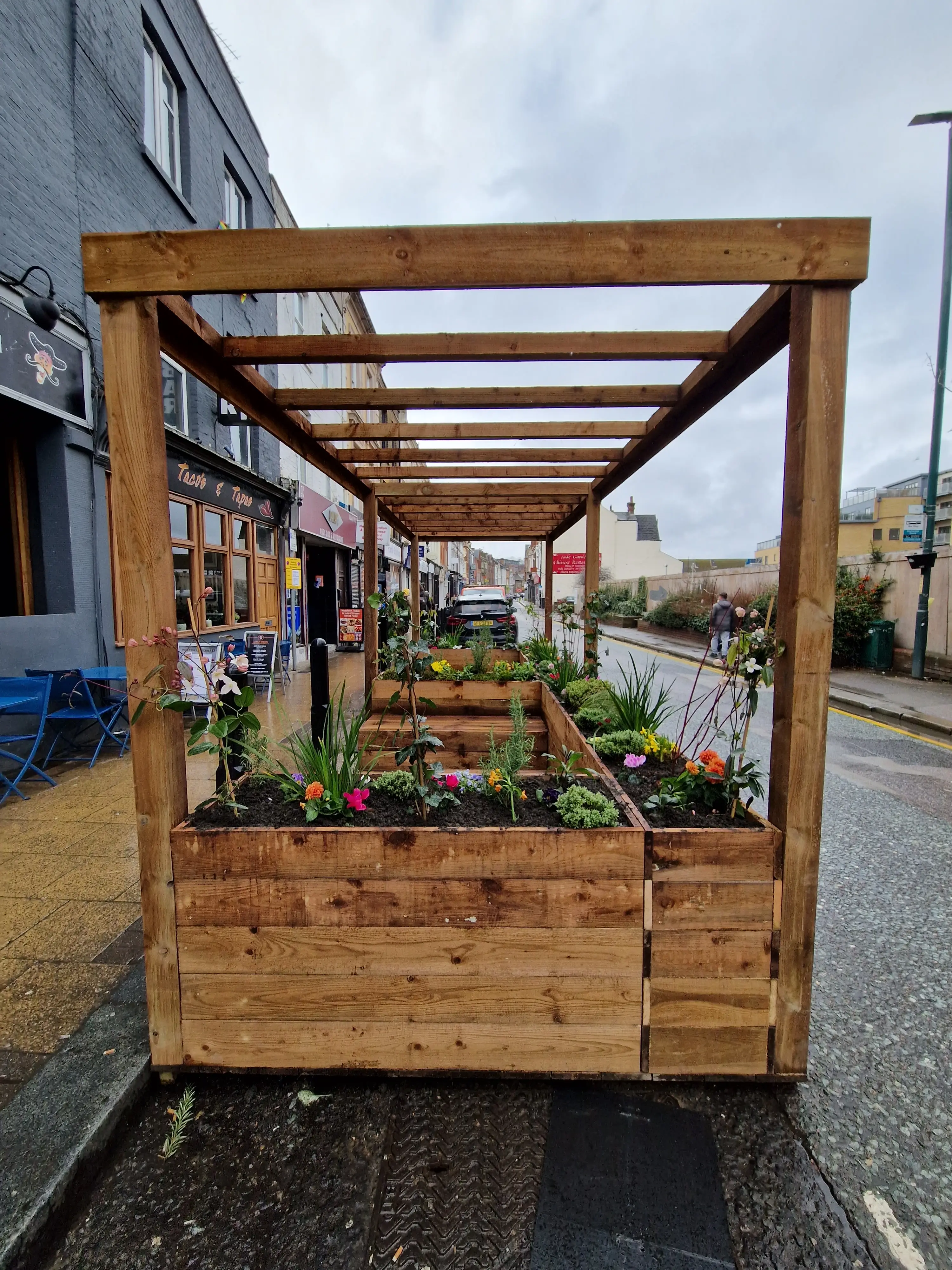 A wooden structure with a roof, holding plants, set on a wet pavement near buildings on a cloudy day.
