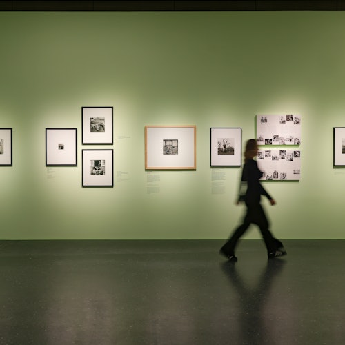 A person walks past a gallery wall displaying framed black-and-white photographs on a light green background.
