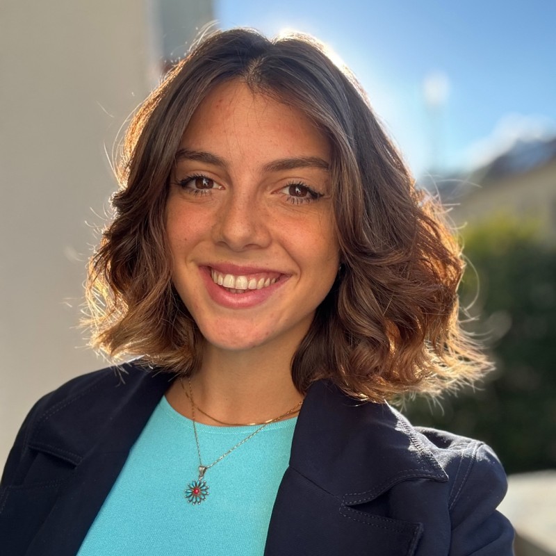 A woman in a white blouse and dark vest sits on a park bench, looking away and smiling. A ukulele rests next to her.