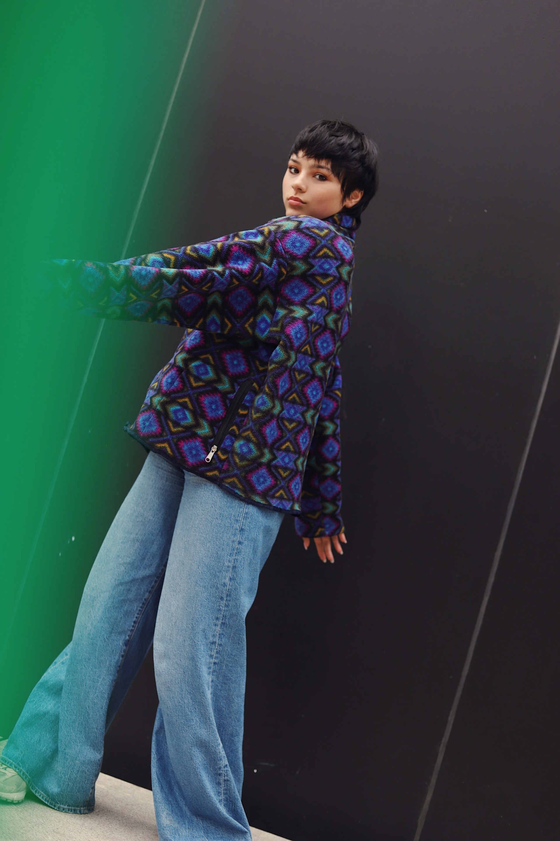 The person in the patterned fleece and wide-leg jeans stands in profile against the same black and green wall, looking over their shoulder toward the camera.