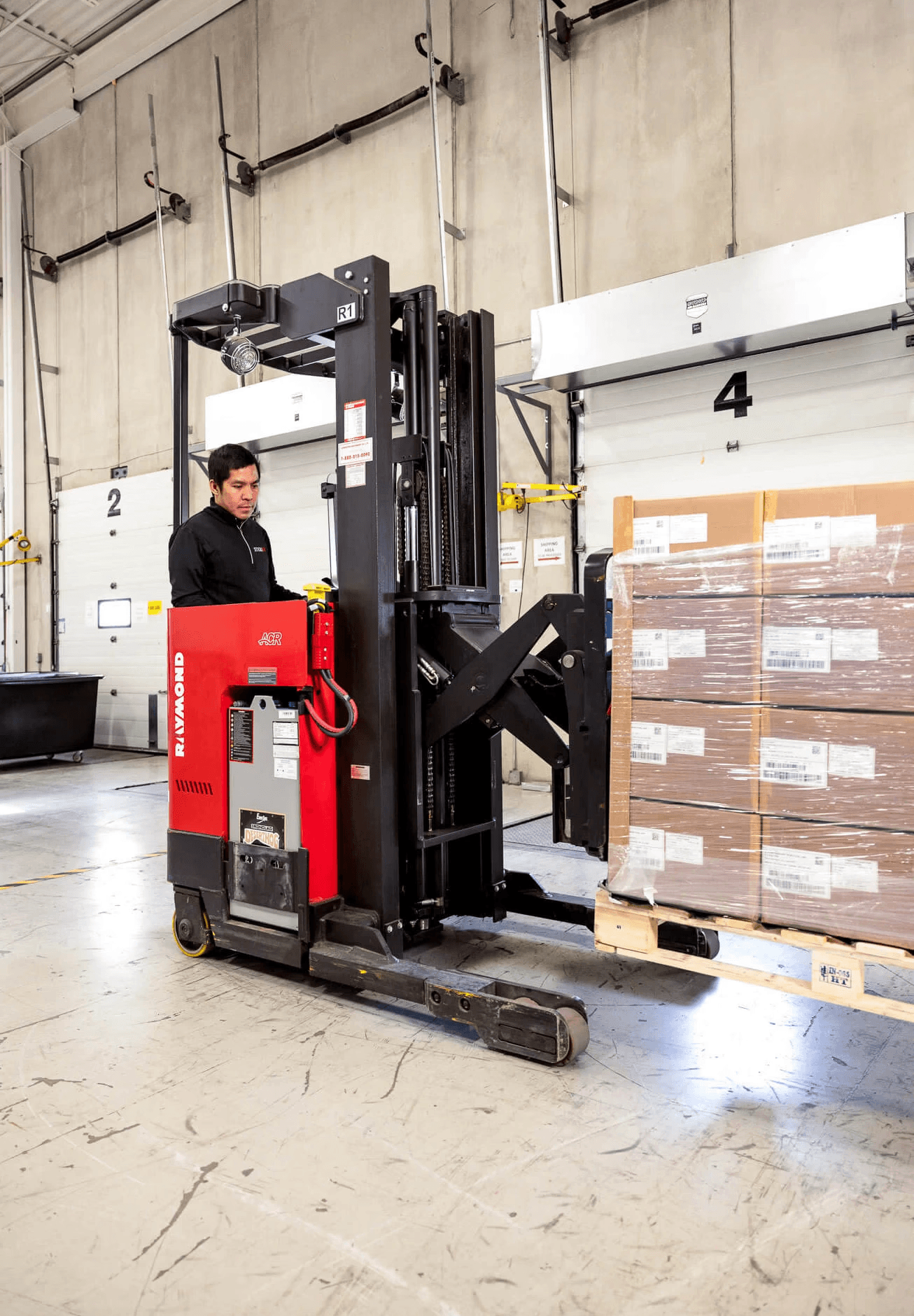 Warehouse worker operates a red forklift to move a wrapped pallet of boxes near loading bay doors.
