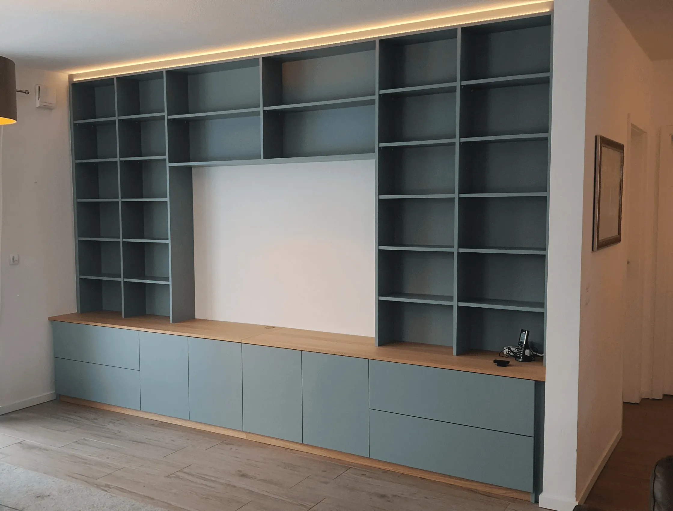 Maßgefertigte Wohnwand in Graublau mit integriertem TV-Bereich, offenen Regalfächern, Schränken und indirekter LED-Beleuchtung.
