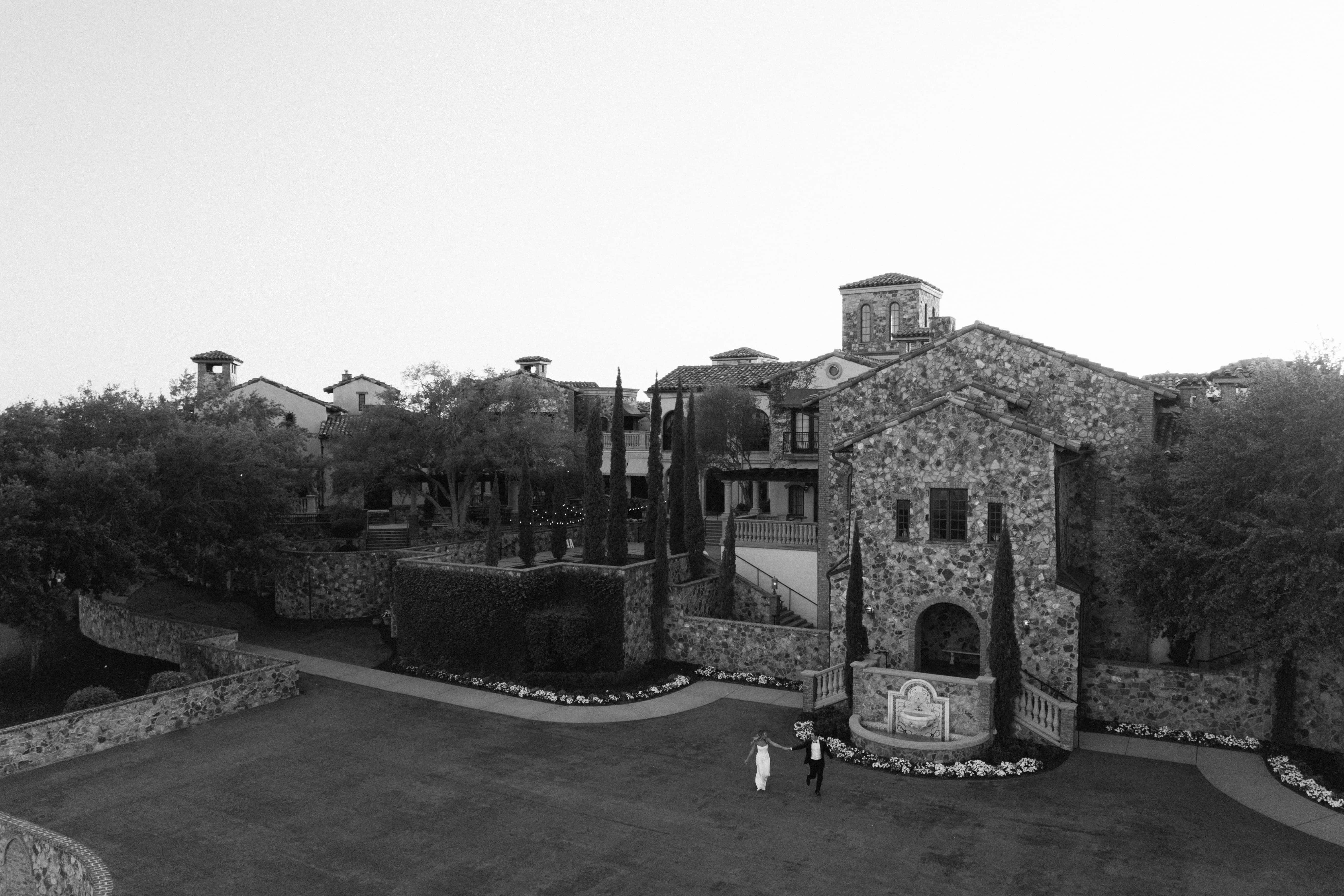  A black-and-white aerial view of a stone building with trees and a large, empty lawn.