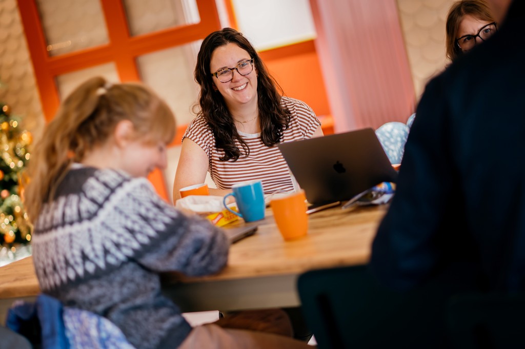 A group of people sit around a table in a warm, bright room. A woman smiles while working on a laptop as others listen, with mugs and notebooks on the table.