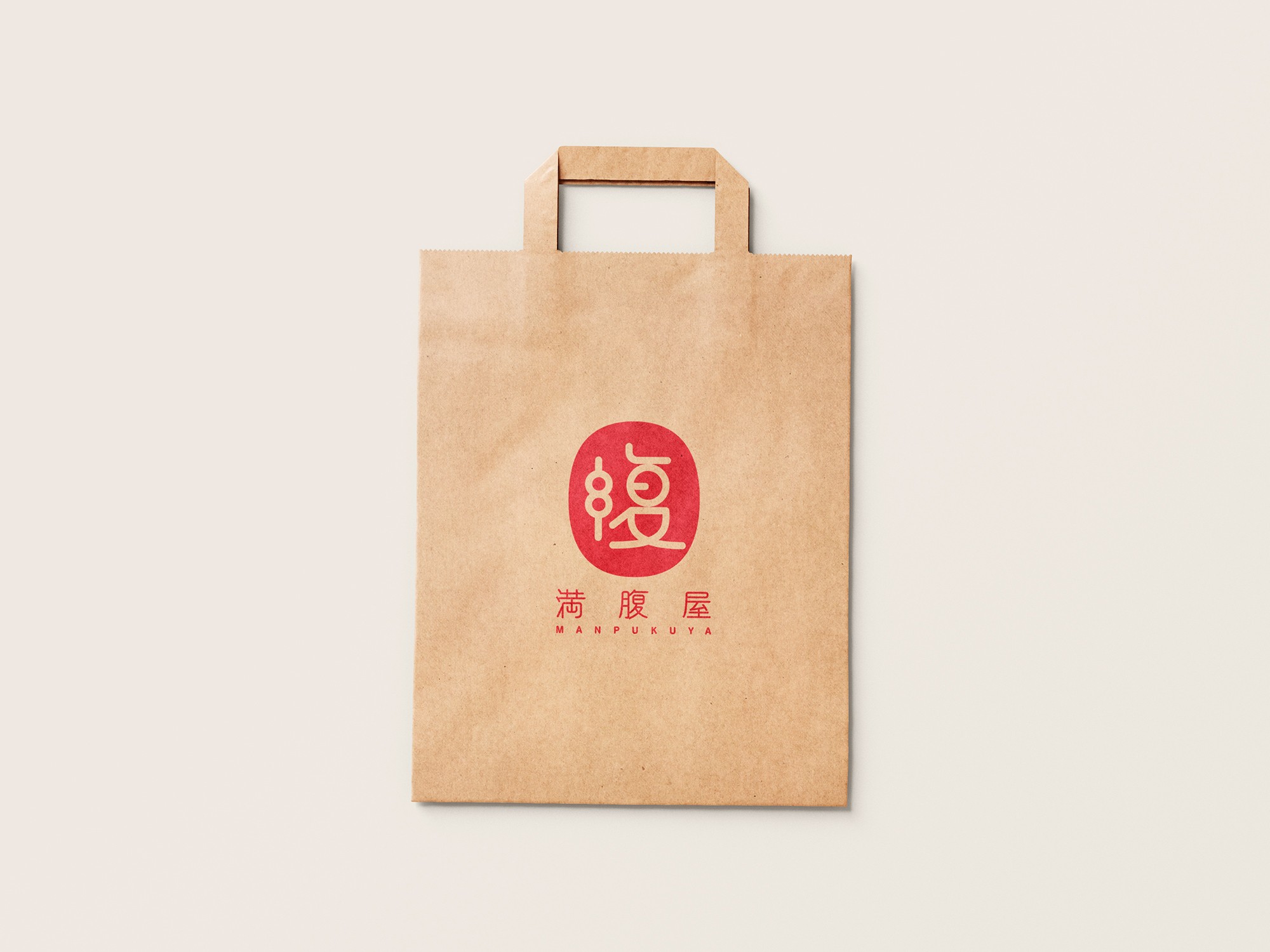 這張圖片展示了滿腹屋的紙袋設計，紙袋正面印有滿腹屋的標識。標識中心有一個紅色圓形內帶有藝術感的白色字「腹」，下方顯示中文「滿腹屋」和英文「MANPUKUYA」。