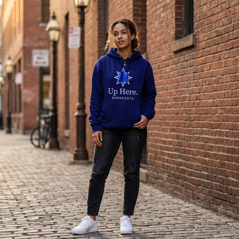 A young multiracial woman stands on an urban brick sidewalk wearing a deep navy Up Here. Minnesota hoodie with the star logo embroidered on the chest.