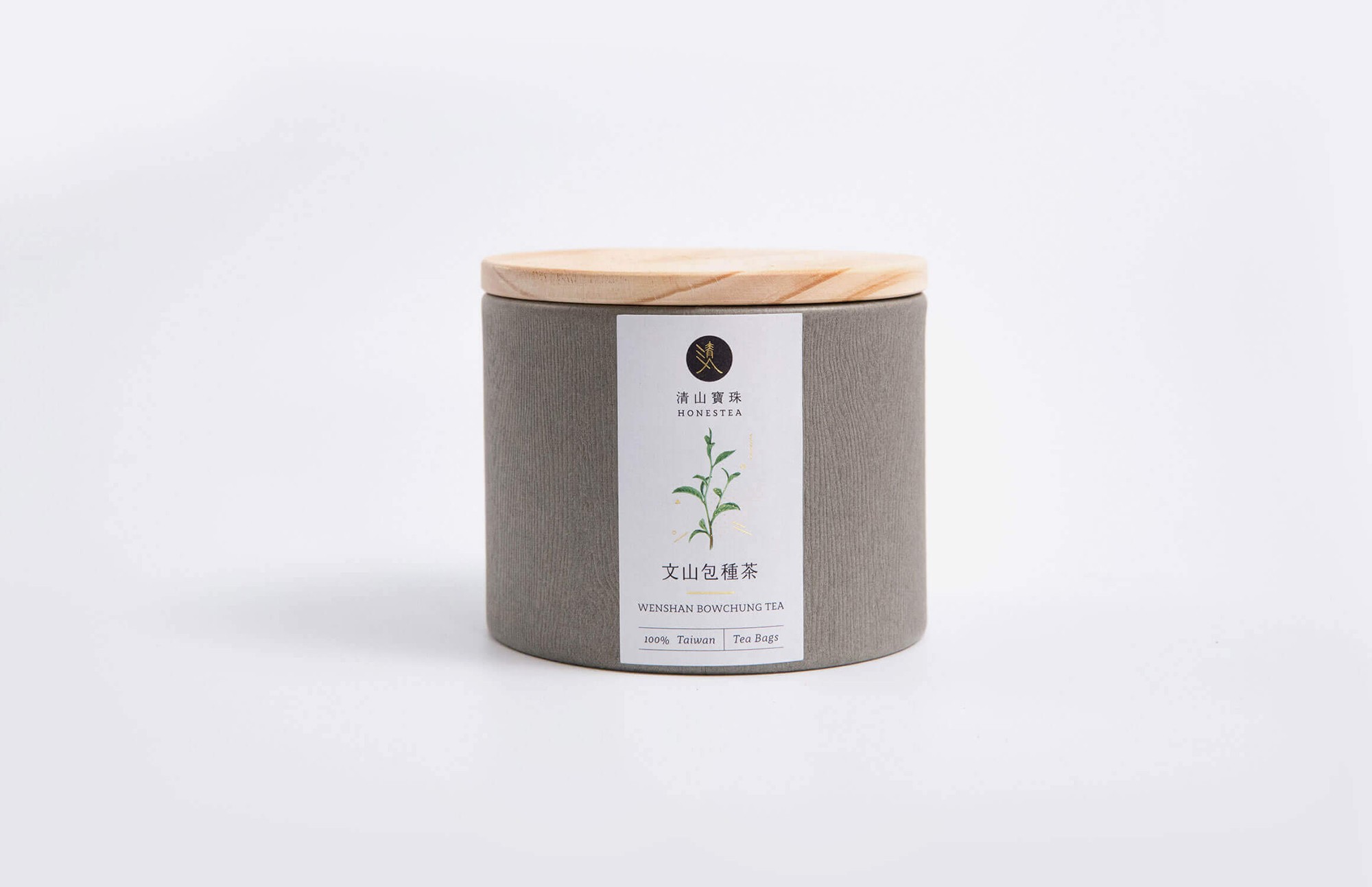 這張圖展示了清山寶珠的文山包種茶圓筒包裝。包裝為灰色紙材，帶有木質蓋子，正面貼有標籤，上印有品牌標誌、一株茶葉的手繪水彩插圖，以及「清山寶珠 HONESTEA」和「文山包種茶」字樣。