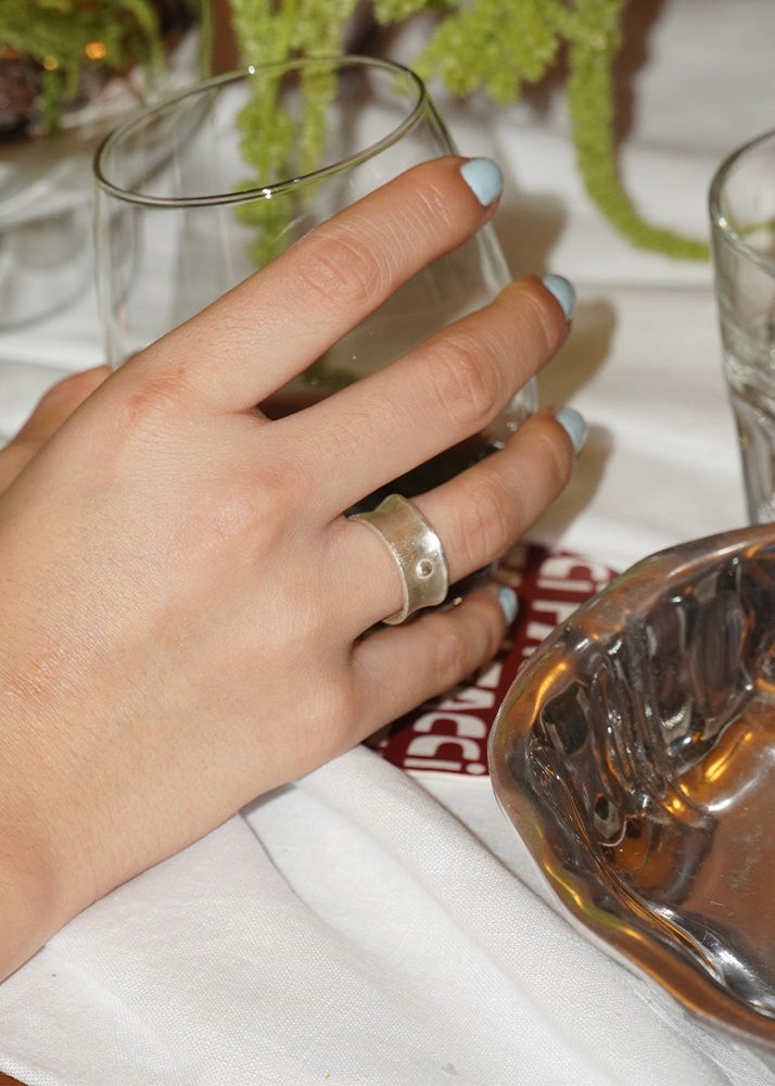 Hand with a silver ring holding a glass on a table with a plant in the background