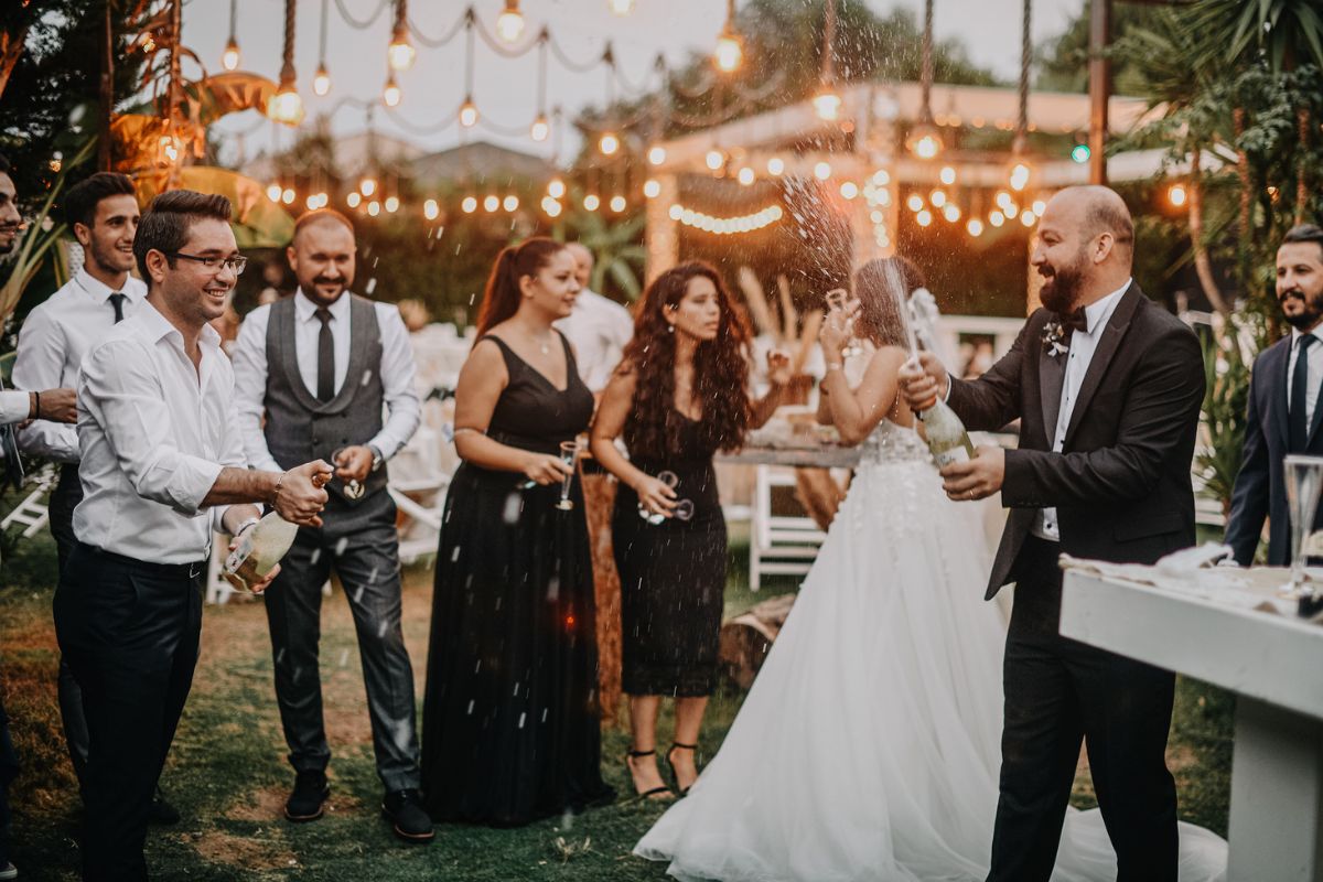 A bride and groom celebrating outdoors, with the groom popping a champagne cork while the wedding party cheers.