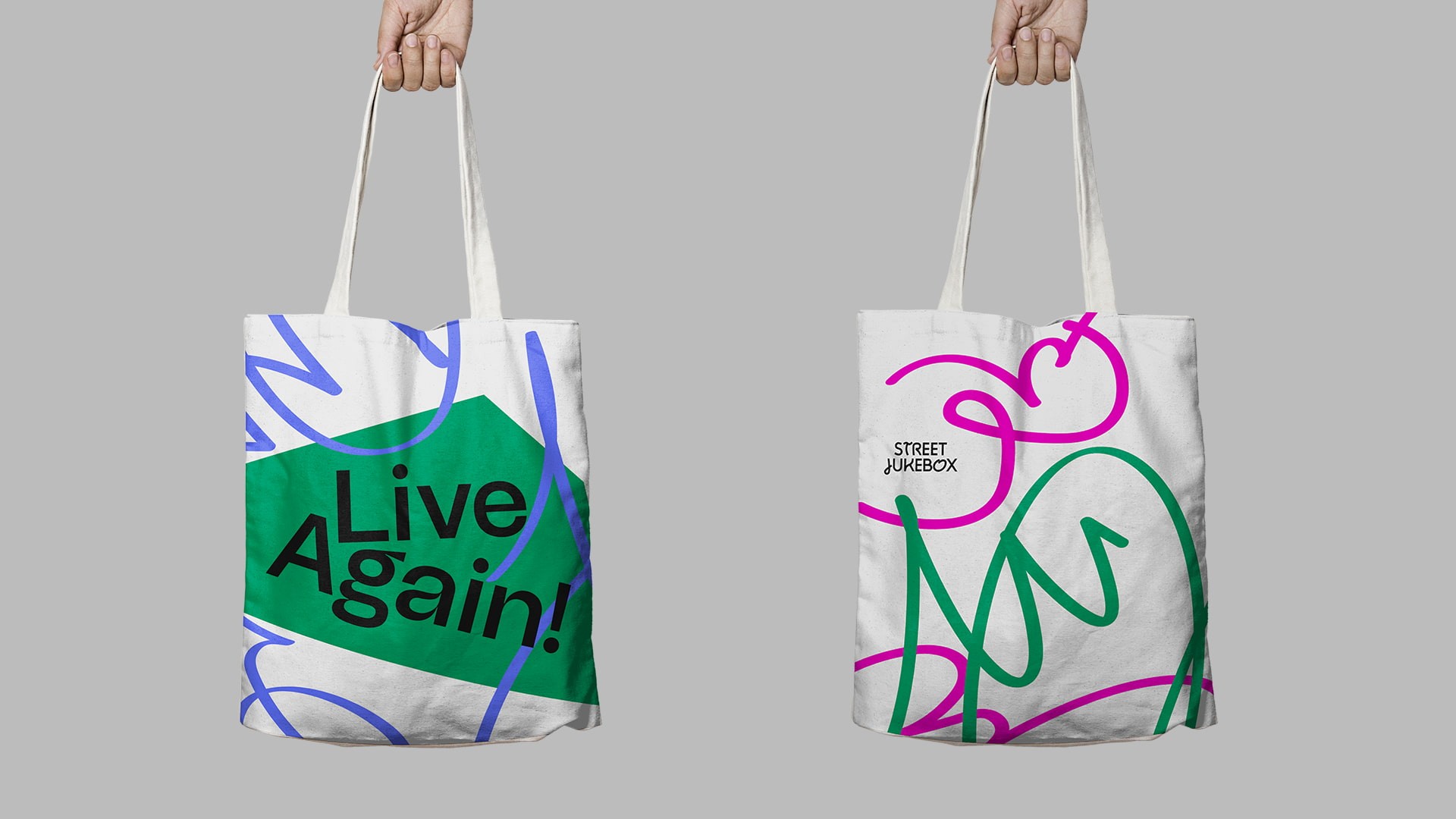 Two white tote bags are held by hands against a gray background. The left bag features a green diamond shape with the words “Live Again!” in black letters. The right bag displays a pink and green abstract design with the words “Street Jukebox” in black text.