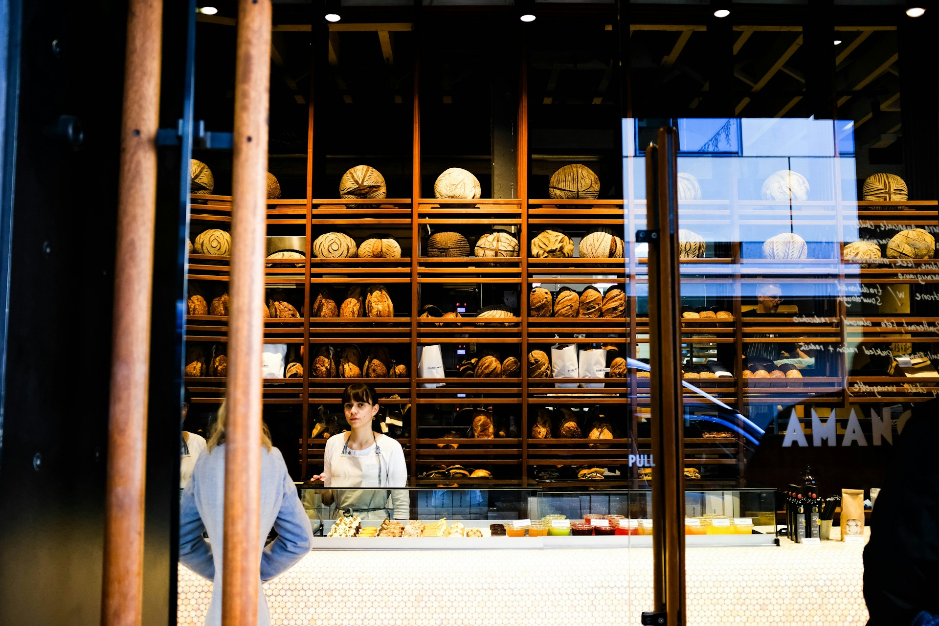 This image shows a woman, possibly a bakery owner or staff member, standing behind a counter filled with pastries and bread in a bakery with shelves of neatly arranged loaves behind her. This image serves as the hero image for the blog on the Rarecide website, discussing 'The Impact of Social Media on Customer Acquisition and Retention.' Rarecide, a top-rated social media management and marketing agency, specializes in social media marketing, marketing strategy development, and customer engagement. Known as the best marketing agency in Toronto, Rarecide excels in innovative marketing strategies for business growth.