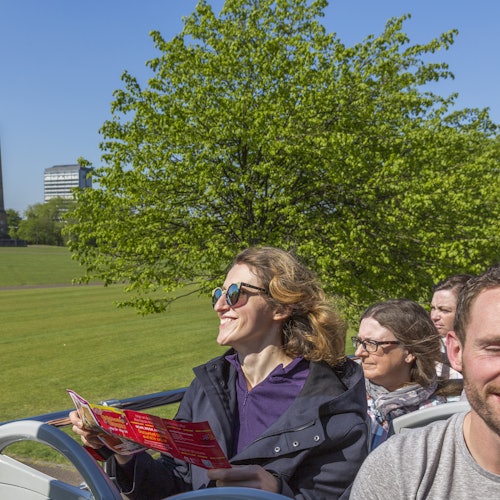 People sitting on a double-decker bus with a grassy park and trees in the background, one holding a pamphlet and smiling.