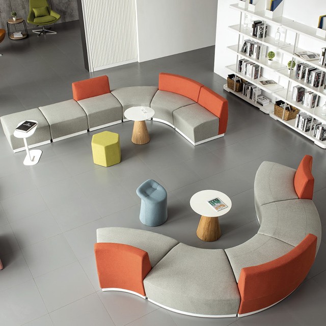 Top-down view of an S-shaped modular office sofa in gray and orange fabric, winding around a large indoor plant with white minimalist side tables and a bookshelf in the background.