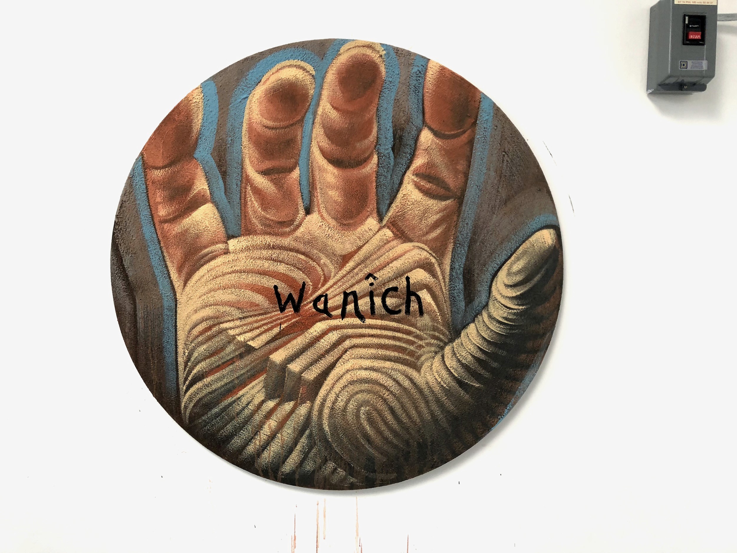 A round painting of an open hand carved with fingerprint-like ridges, the word “Wanîch” written on the palm.