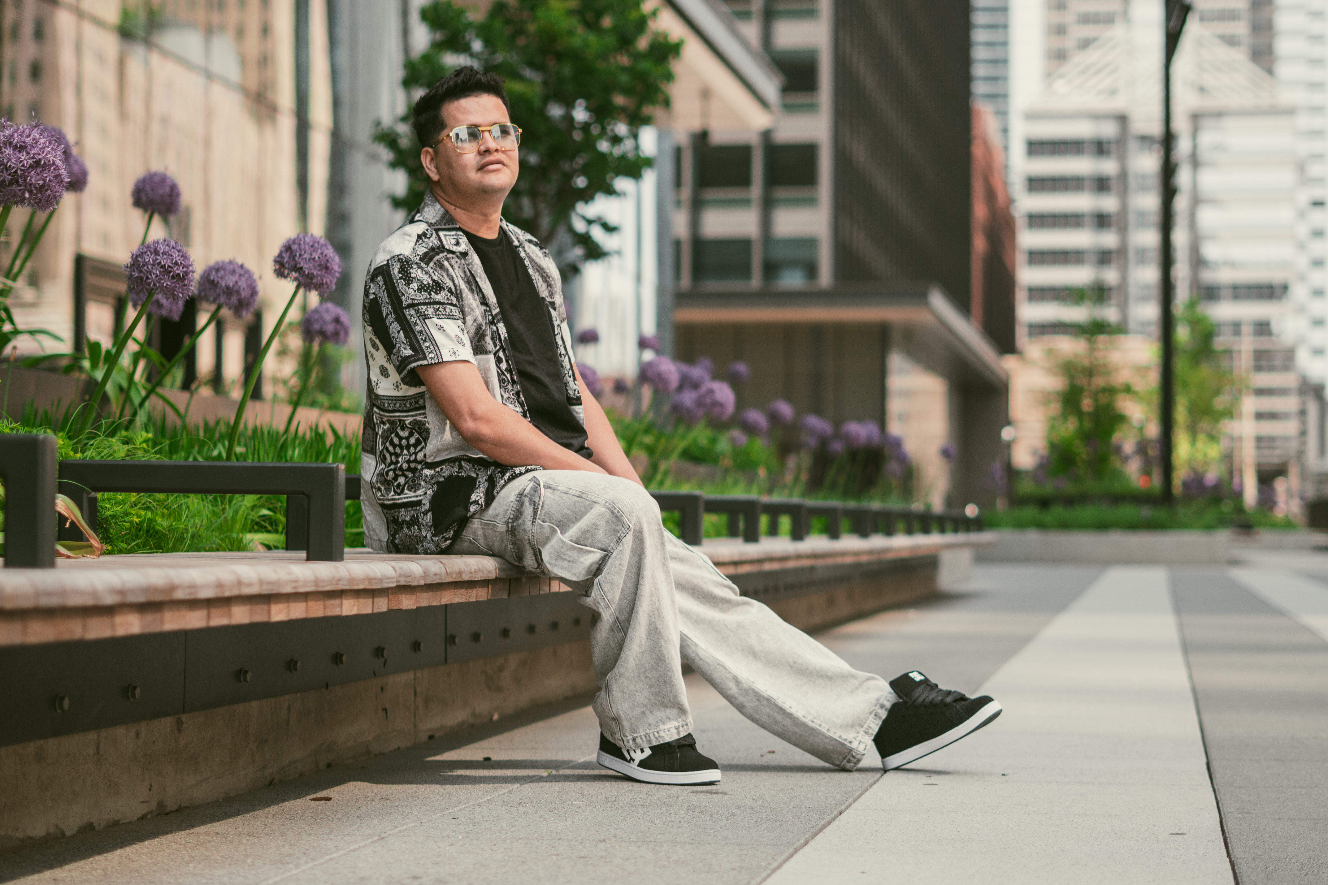 Dani Sierra sitting casually on a low wall, surrounded by greenery and urban buildings in the background.
