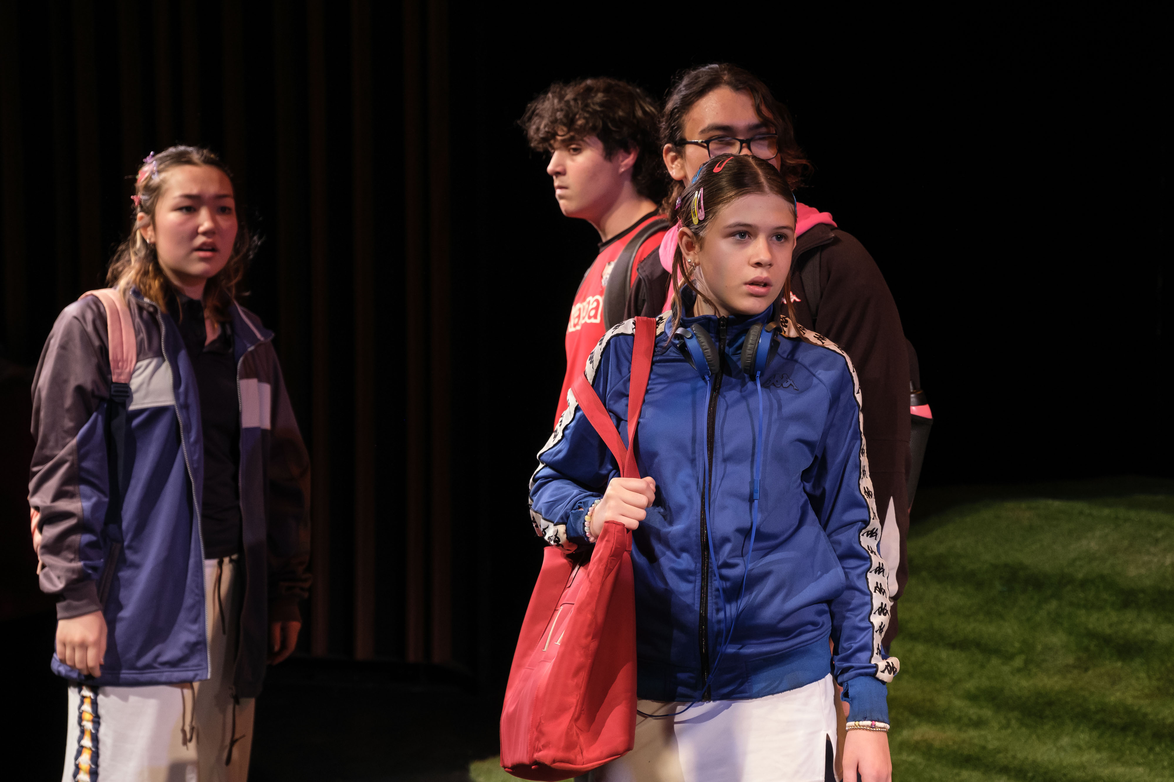A small group of students stands close together, looking offstage with concerned expressions. The lead performer in a blue tracksuit grips a red bag, suggesting a tense or uncertain moment in the narrative.