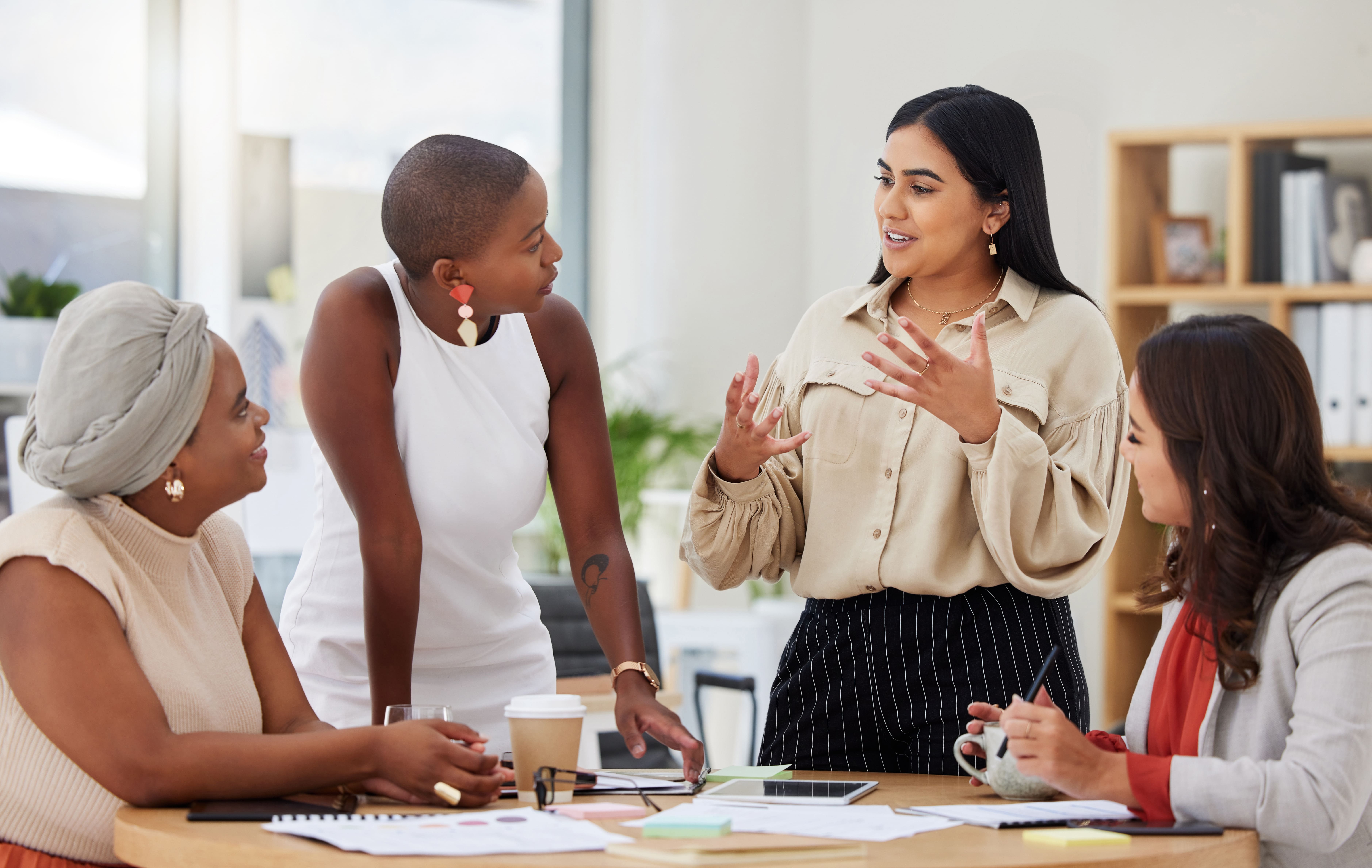 Imagem de um grupo diverso de mulheres reunidas em uma mesa, participando de uma conversa ou discussão de trabalho. Uma das mulheres, de pé, gesticula enquanto fala, atraindo a atenção das demais, que escutam atentas. A atmosfera parece colaborativa e acolhedora, evidenciada pelas expressões engajadas e a linguagem corporal descontraída. Ao fundo, estantes e plantas complementam o ambiente, sugerindo um escritório moderno e inclusivo, onde a diversidade e a representatividade feminina são celebradas.