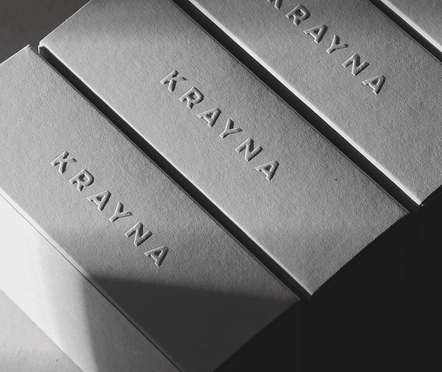 A diagonal stack of white Krayna boxes with embossed logos, captured in dramatic light and shadow. This visual reinforces a minimalist brand architecture and high-end positioning suitable for global distribution.