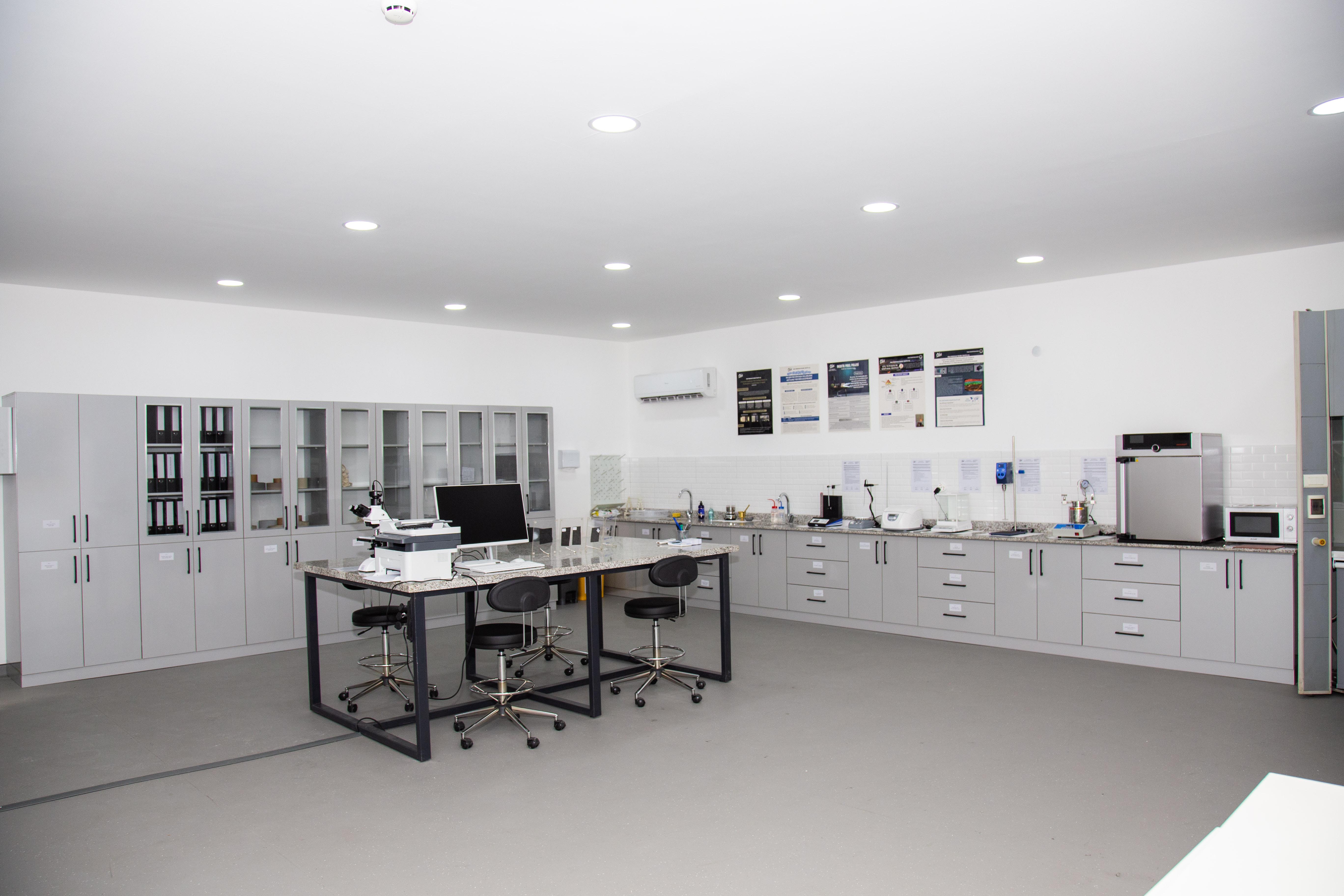Two men sit at a wooden table with laptops, under a large red lamp. A woman walks by a window. The scene is calm, set against a red-brown wall.