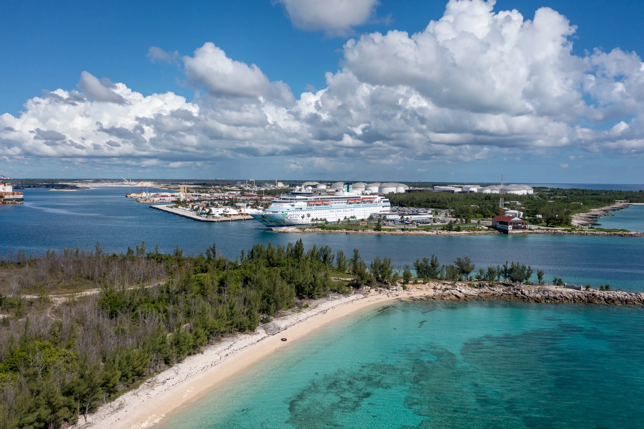 Aerial view of a cruise ship docked at a port, surrounded by blue water and green trees, with a sandy beach in the foreground and partly cloudy sky above.