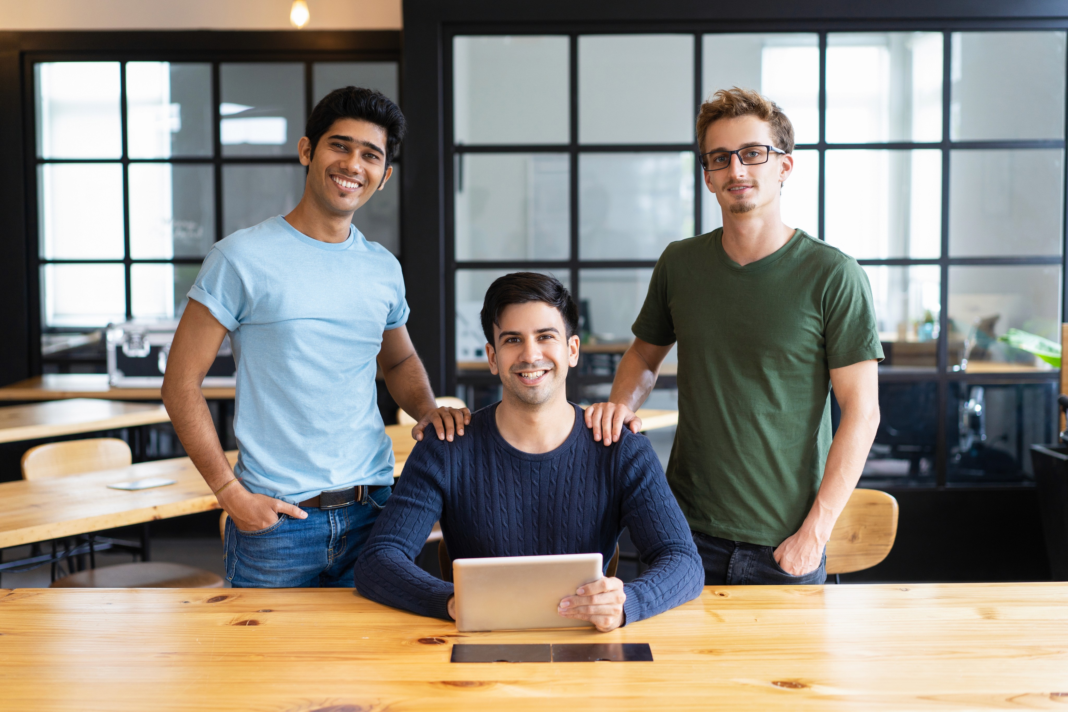 Three young men in a modern office, with one seated holding a tablet and two standing beside him.